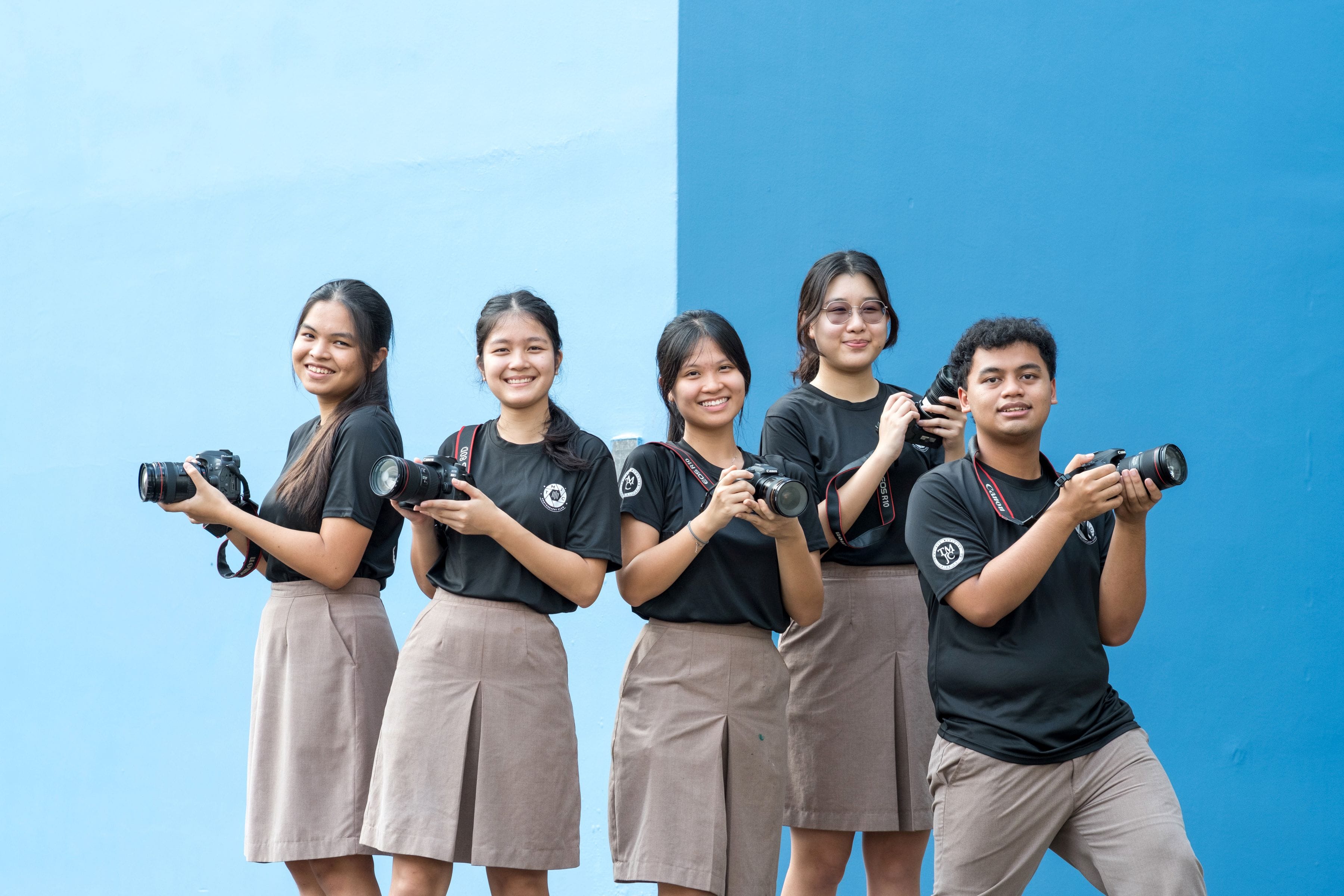 Five smiling students in black TMJC club T-shirts and khaki bottoms stand in a row against a two-tone blue wall. Each student holds up a Canon DSLR camera. They are posed at a slight angle, looking towards the camera.